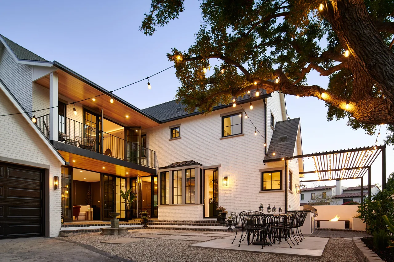 Outdoor evening view of a white brick two-story home with a balcony and an outdoor dining space under string lights.