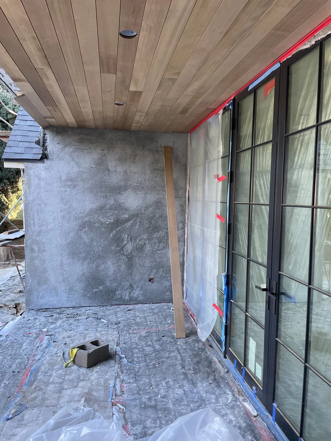 View of a covered patio area under construction with wood plank ceiling and glass doors protected by plastic sheeting.