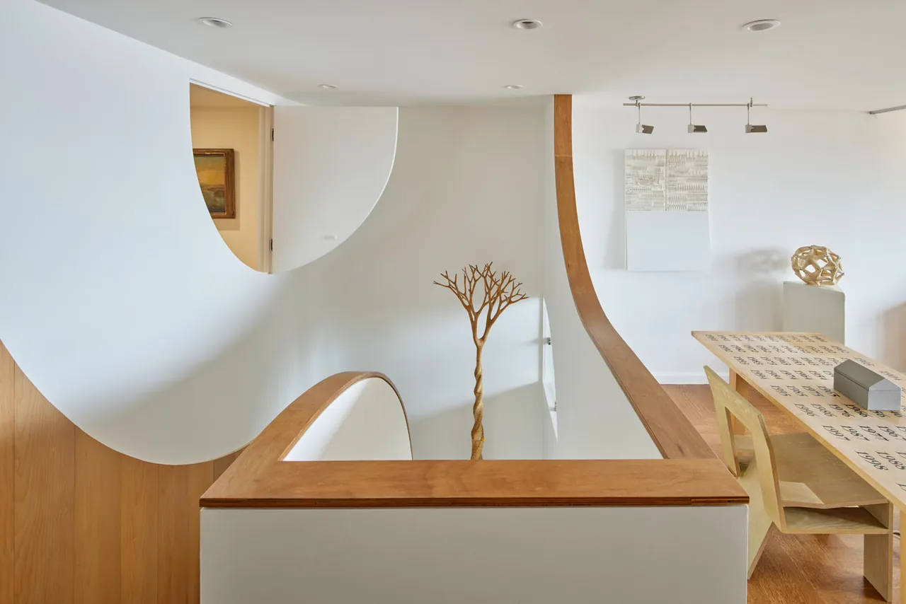 Curved wood and white walls define a modern stairwell with a decorative wooden tree sculpture and a nearby dining table.