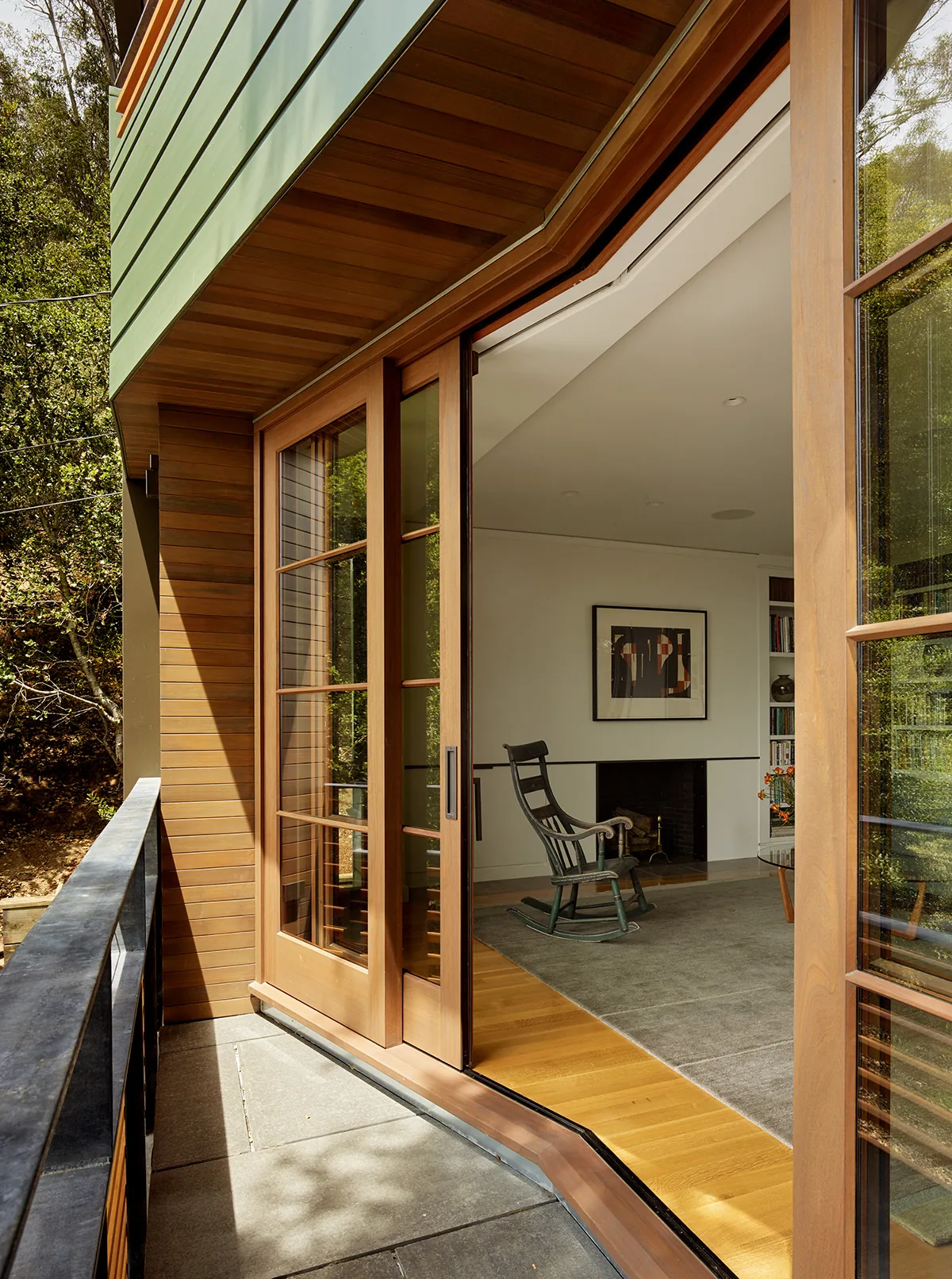 Wood-framed sliding glass doors open from outdoor balcony into a living room featuring a fireplace and wooden rocking chair.