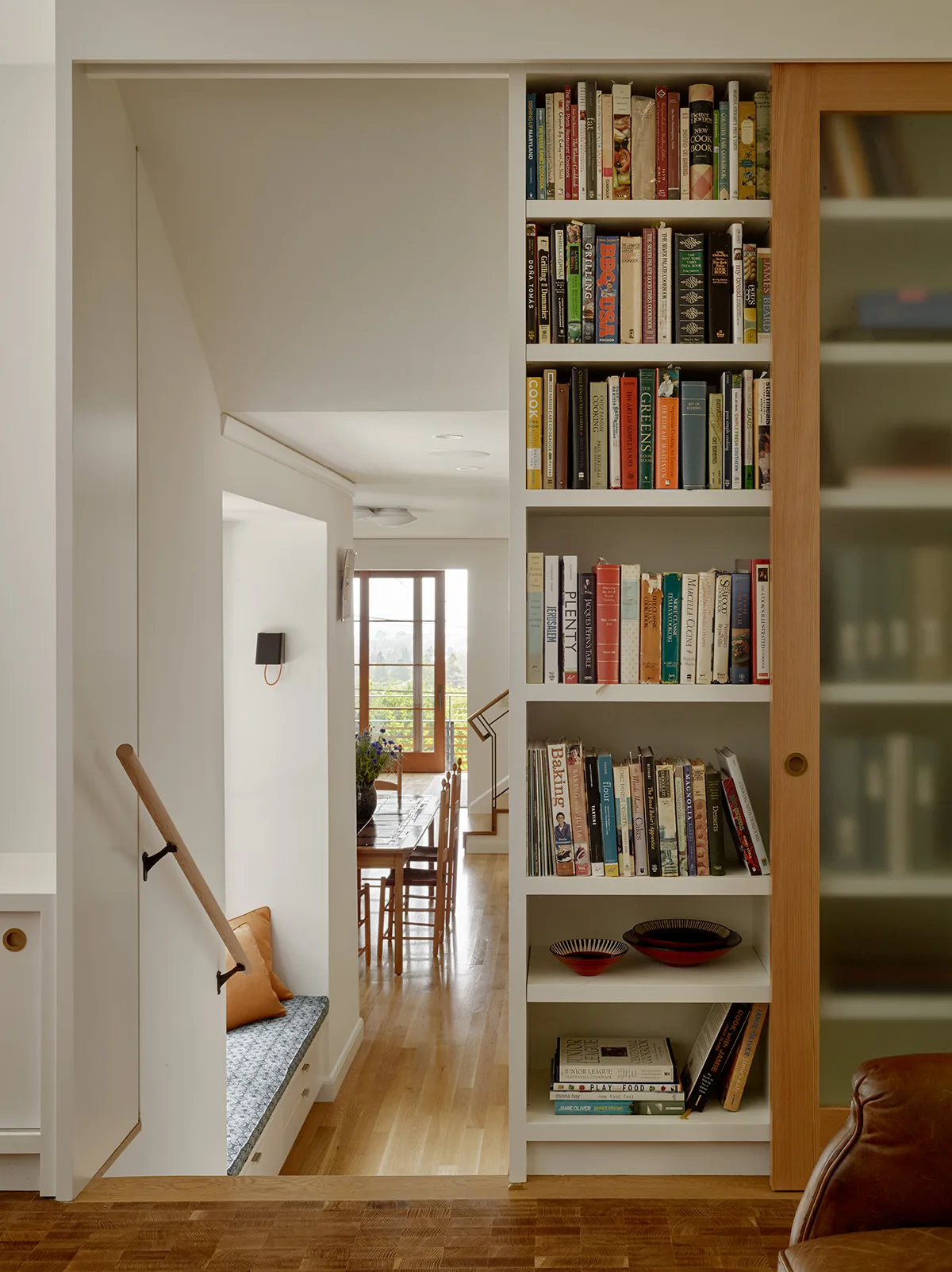 Built-in bookshelf with sliding wooden door and window bench in hallway opening to dining area with hardwood flooring.