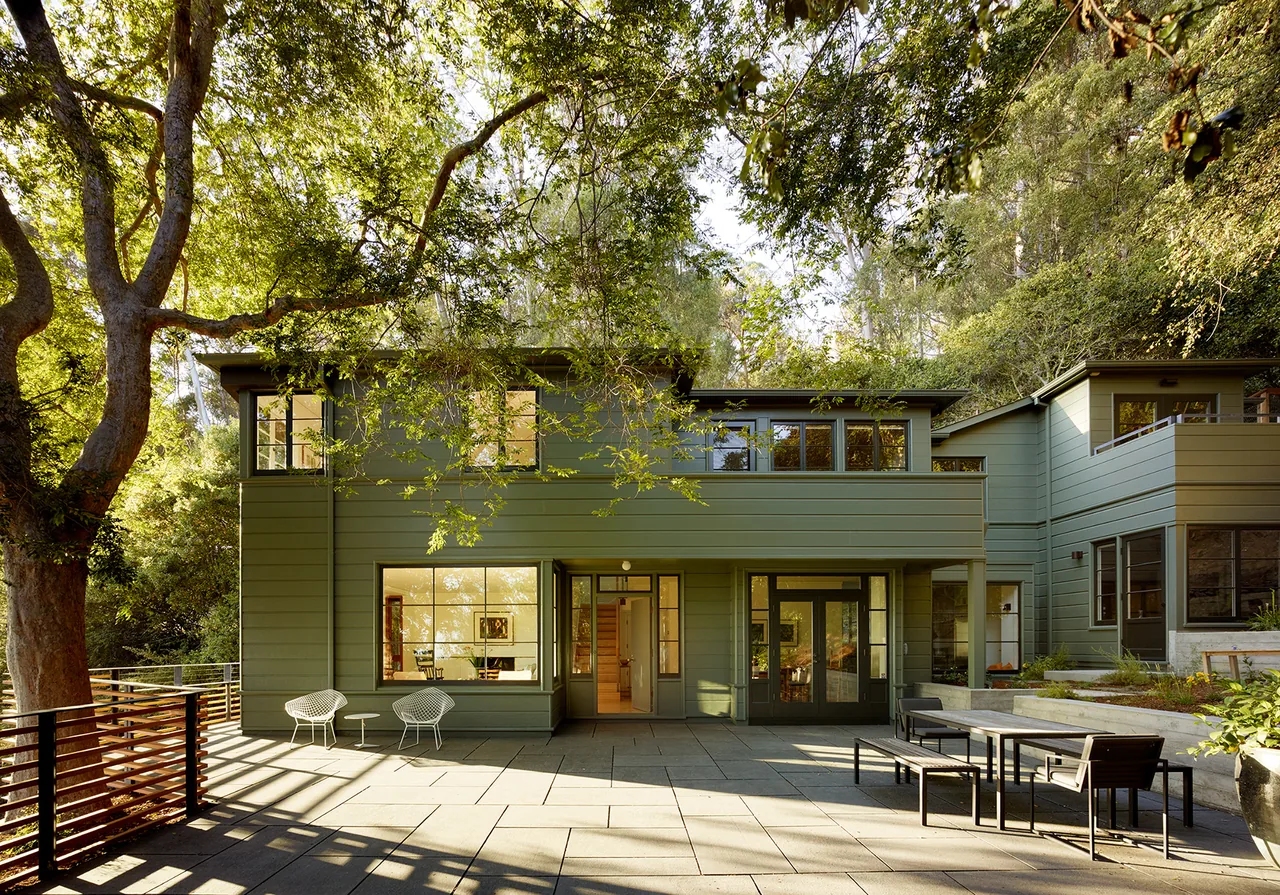 The Claremont Canyon Residence features green wood siding and large windows overlooking a stone patio under mature trees.