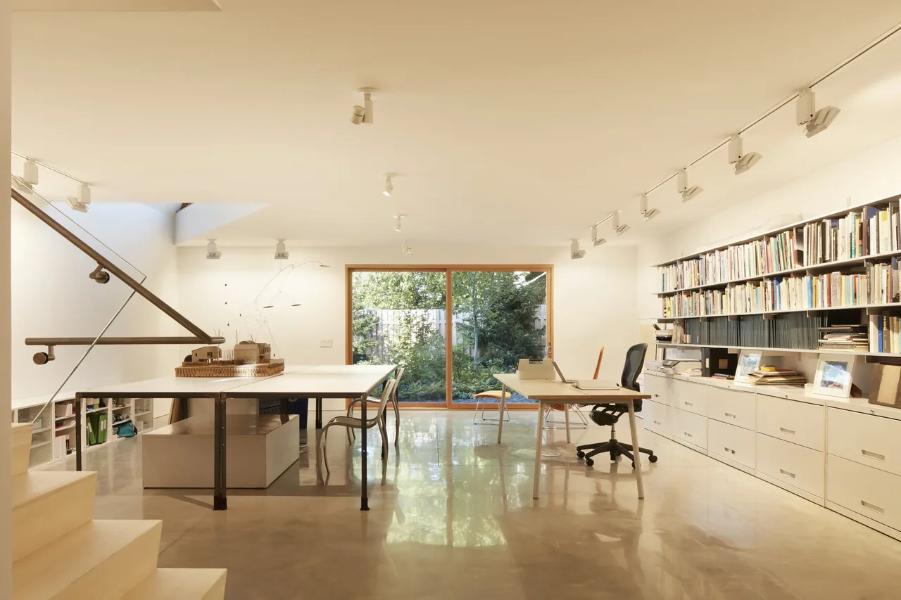 Modern home office features large glass doors with garden view, polished concrete floors, and built-in shelving with books.
