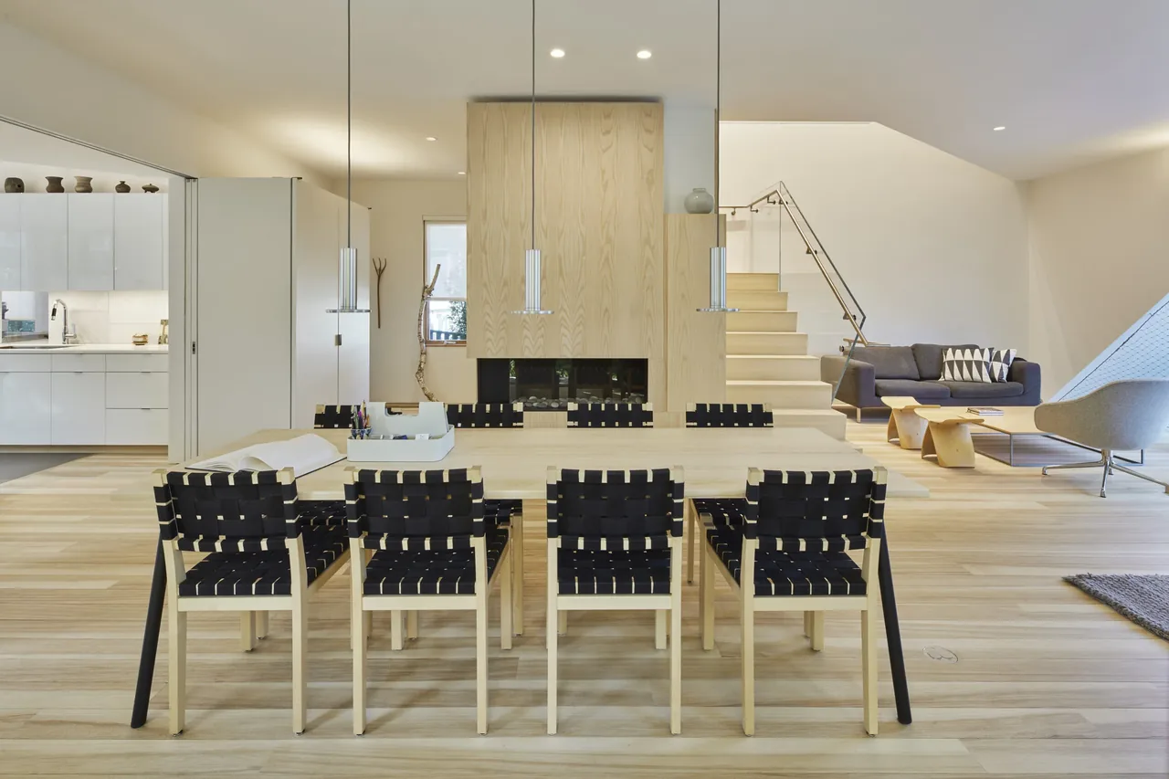 Open floor plan shows a dining table with woven strap chairs, a wood-paneled fireplace, and a living room with modern furniture.