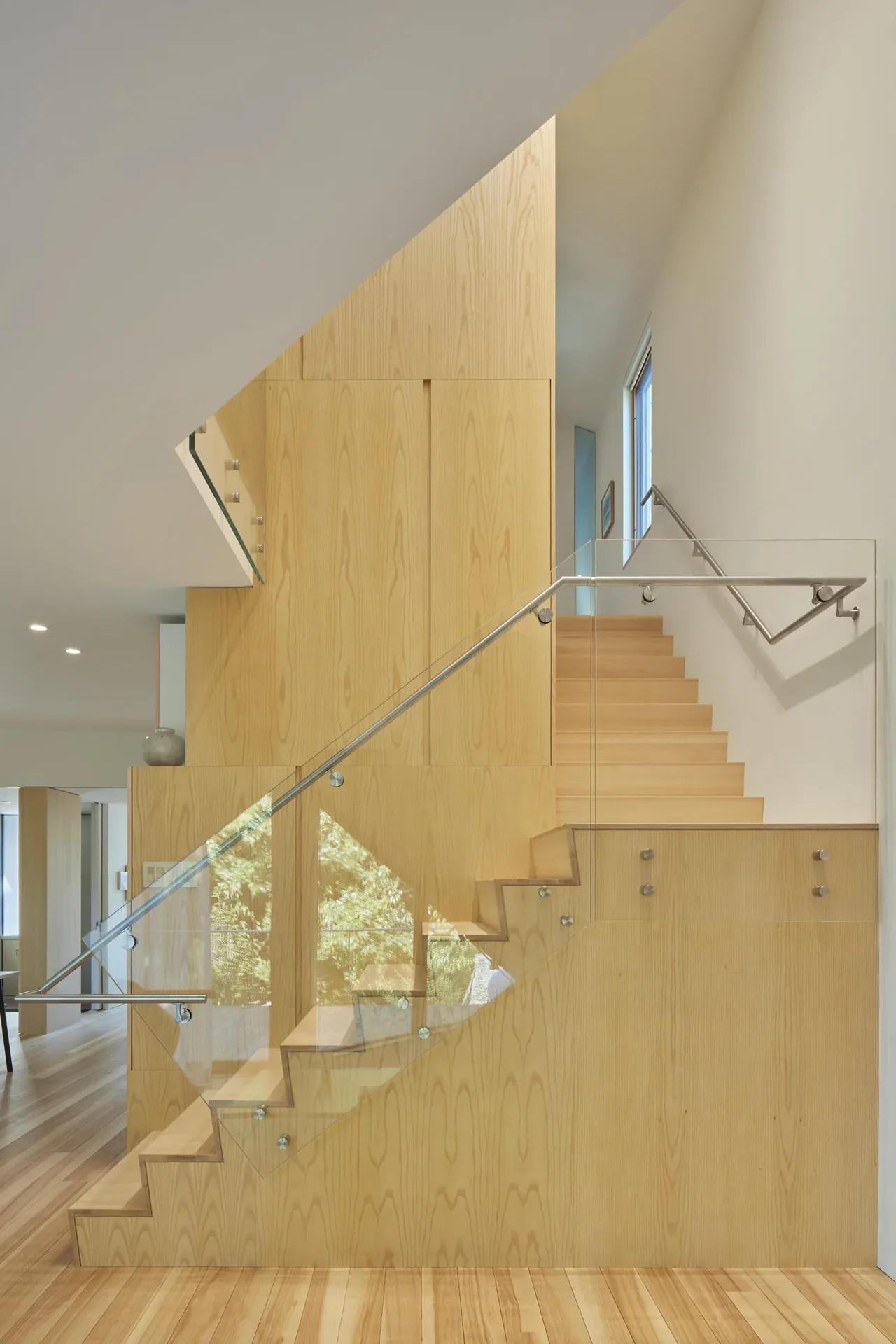 Light wood staircase with glass railing and stainless steel handrails in a bright interior space.