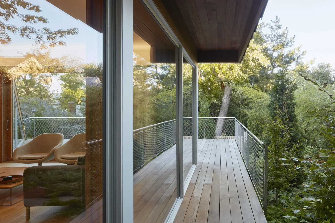A wood-planked balcony extends along the side of the house, bordered by a metal mesh railing, overlooking dense greenery.