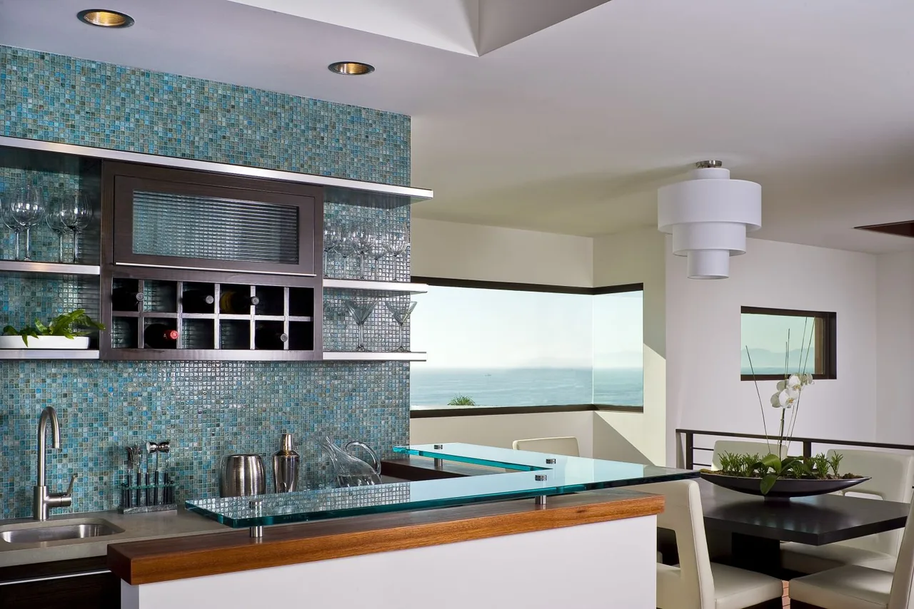 Kitchen bar area with blue mosaic tile backsplash, glass shelving, and corner windows showing an ocean view.