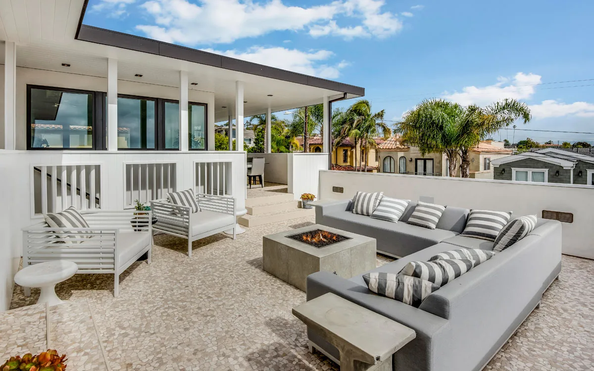 Outdoor patio seating area with gray sectional sofa, white chairs, and a central fire pit under a partly covered roof.