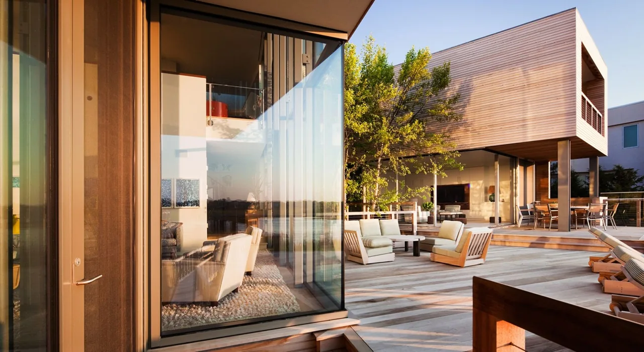 Bay House features a large outdoor deck with wood siding and floor-to-ceiling glazing connecting interior and exterior spaces.