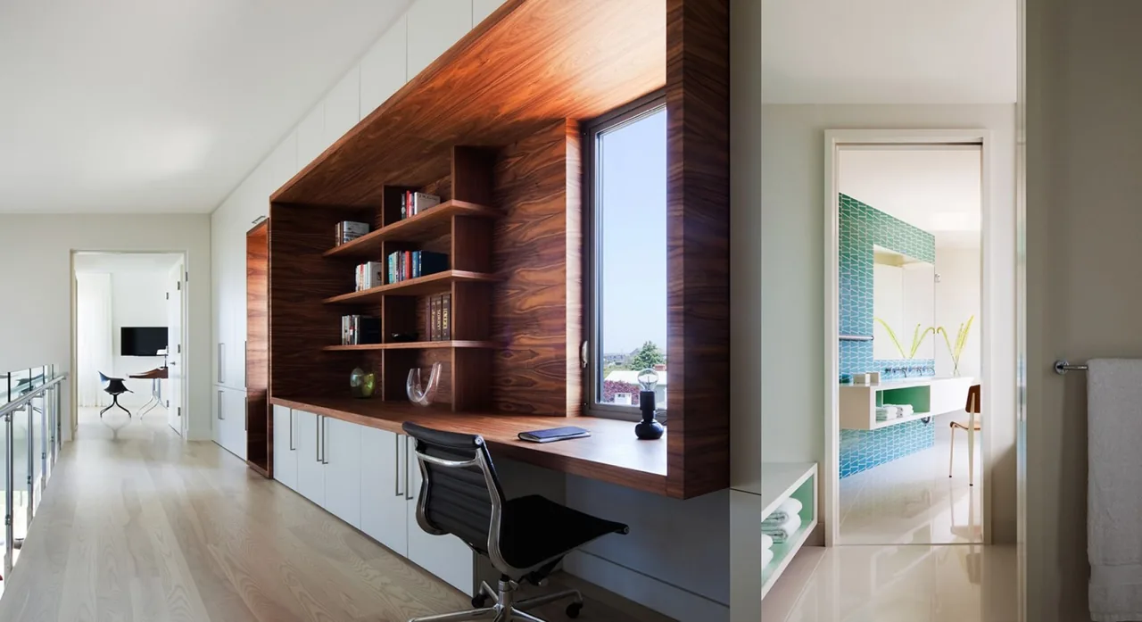A hallway workspace with walnut built-in shelving and desk around a window, revealing a blue-tiled bathroom and a living room beyond.