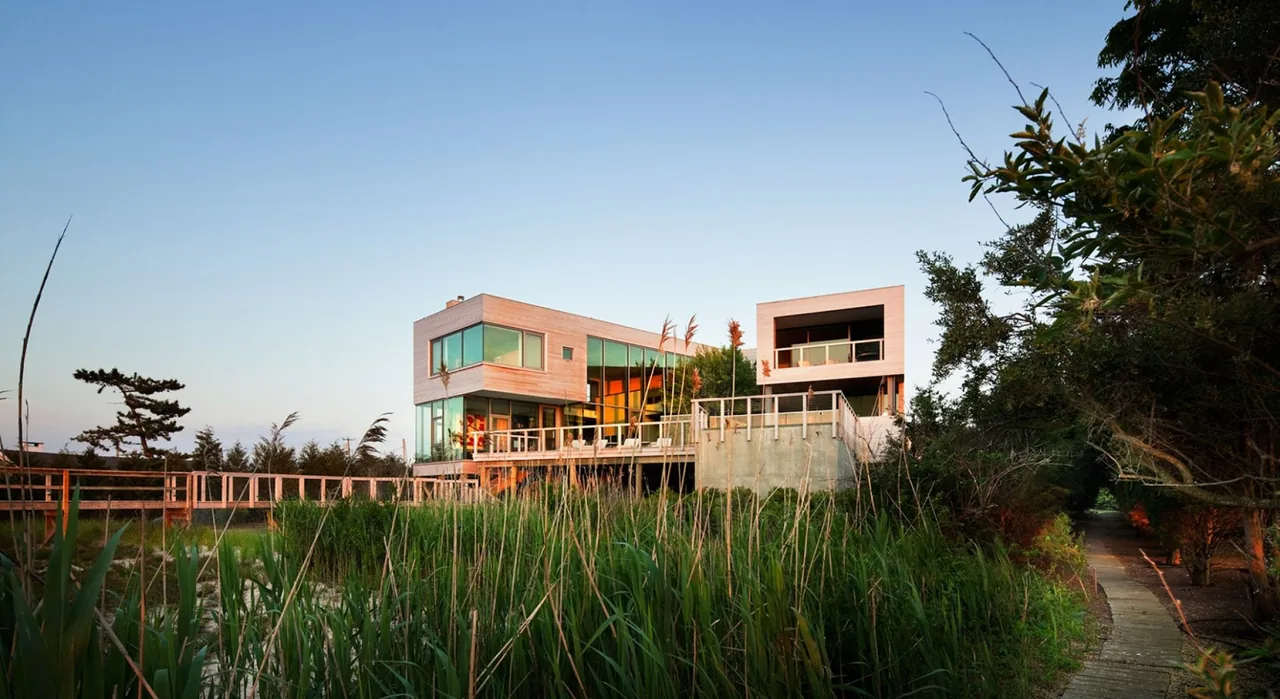Bay House by Leroy Street Studio features large windows, wood siding, and a walkway over wetland vegetation, evoking a modern style with waterfront views.