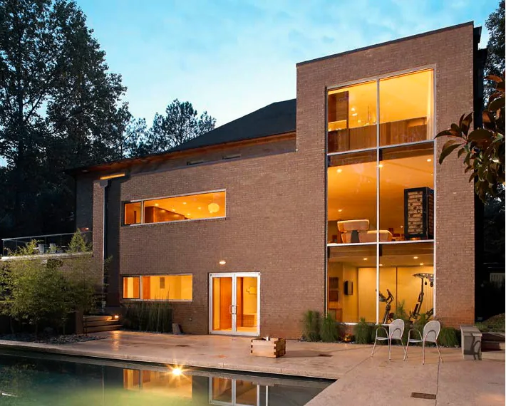 A modern two-story brick house exterior with large vertical windows and an illuminated interior at dusk beside a swimming pool.