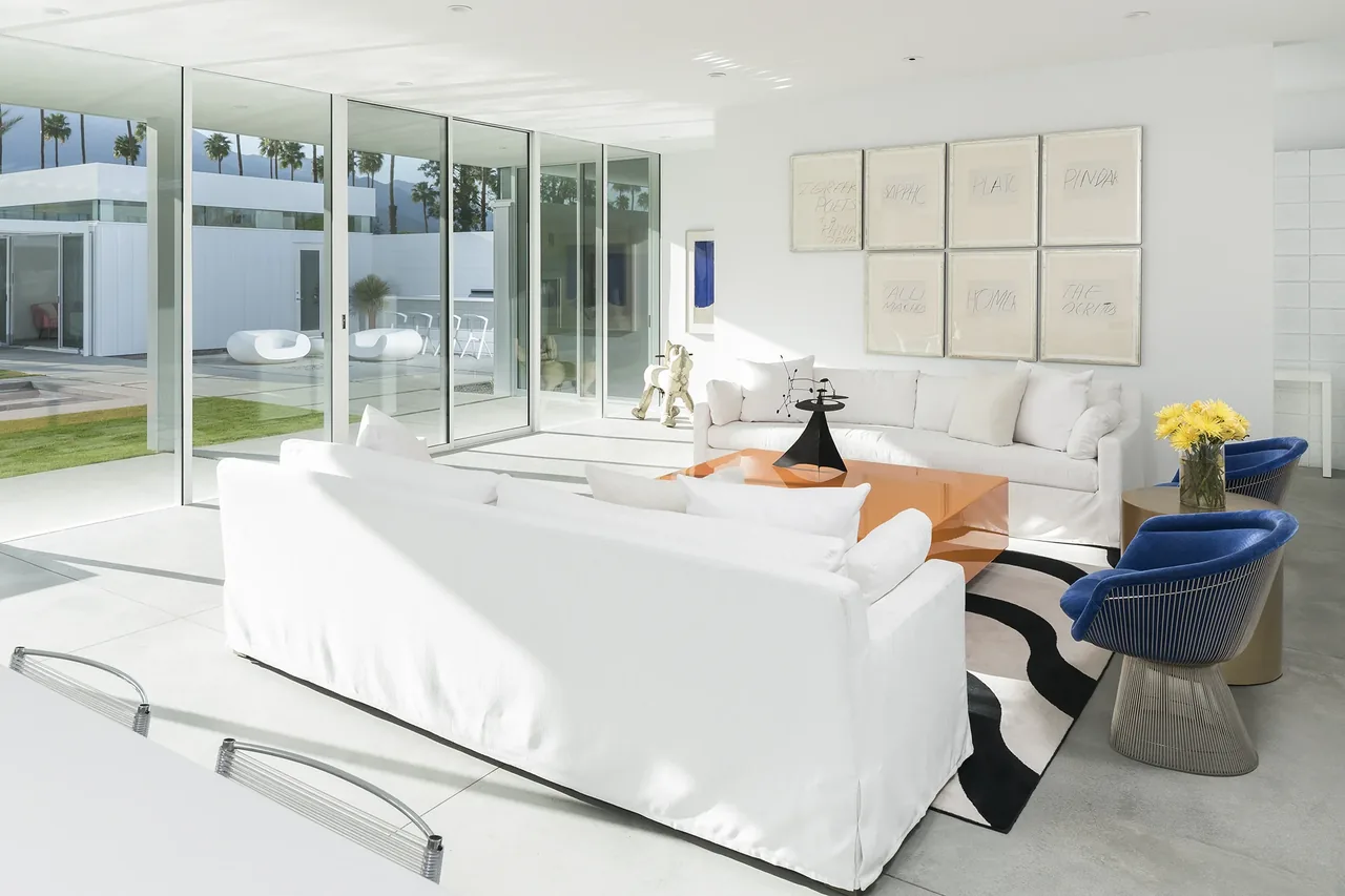 Living room with white sofas, blue accent chairs, and floor-to-ceiling glass walls looking out to an outdoor seating area.