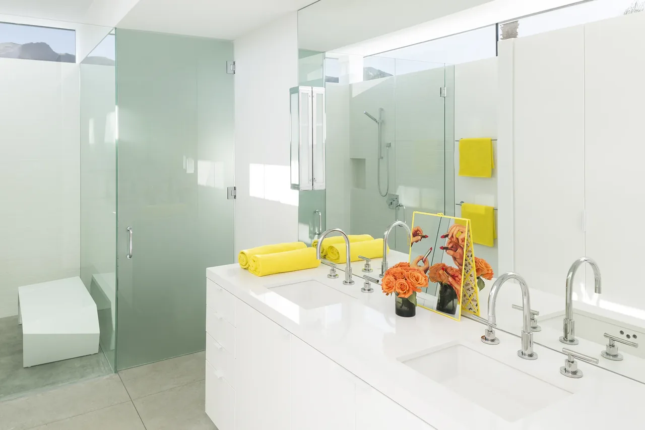 Minimalist white bathroom featuring a double vanity with chrome faucets, frosted glass shower enclosure, and yellow towel accents.