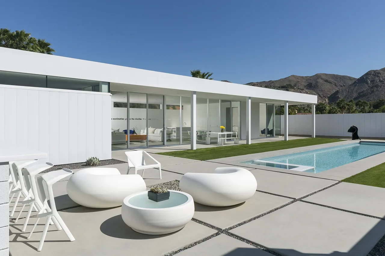 PALM SPRINGS RESIDENCE features a rectangular pool and white modern furnishings outside, with floor-to-ceiling glazing on the house exterior.