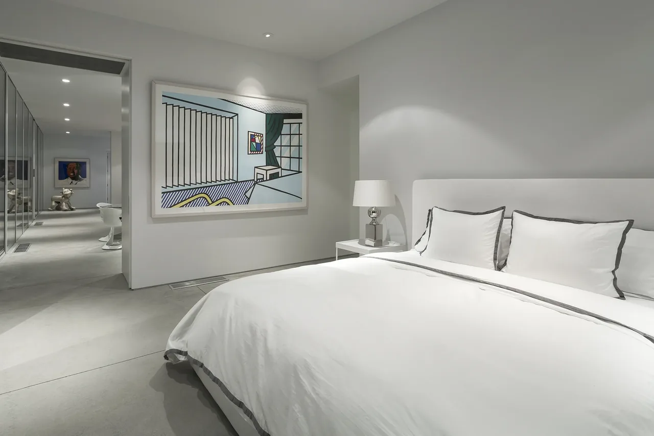 Modern bedroom with white bedding, bedside lamp, and a large piece of contemporary artwork on the wall.