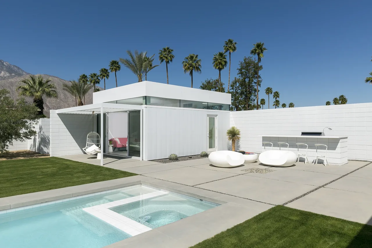 Minimalist pool house with outdoor lounge and bar area next to a rectangular pool, set in a desert landscape with palm trees.