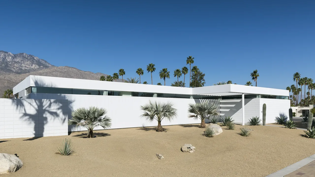 Modern low-profile residence with white exterior walls and desert landscaping featuring palm trees and cacti.