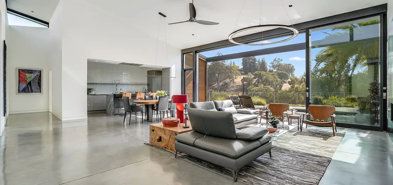 Modern living room features large glazing walls, polished concrete floors, and integrated dining and kitchen spaces.