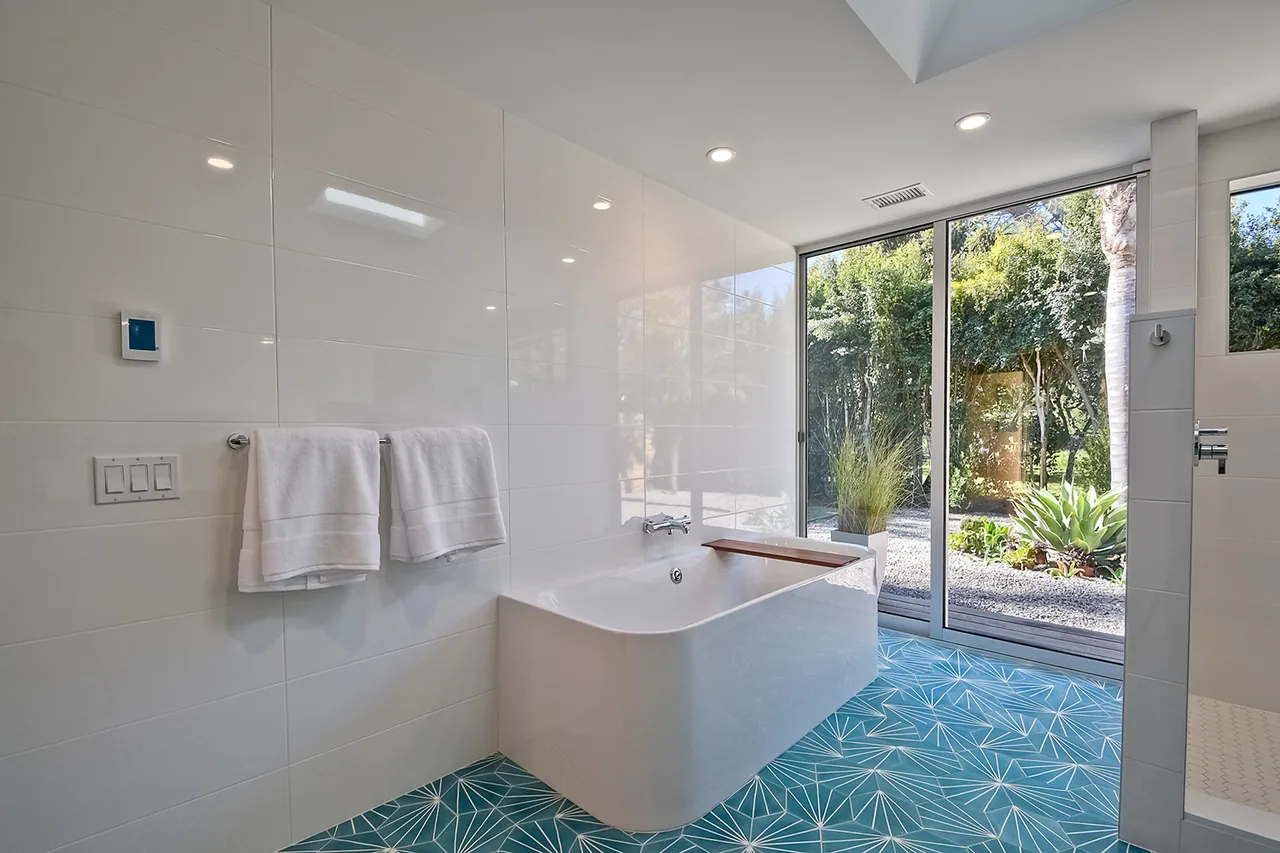 Bathroom with white tile walls, geometric blue floor tiles, and a freestanding tub facing a large glass door to the garden.