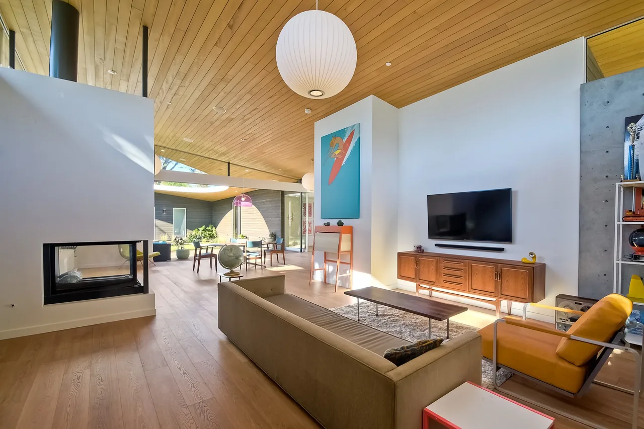 Aa House living and dining space features a wooden ceiling overhang, midcentury modern furniture, and a double-sided fireplace.