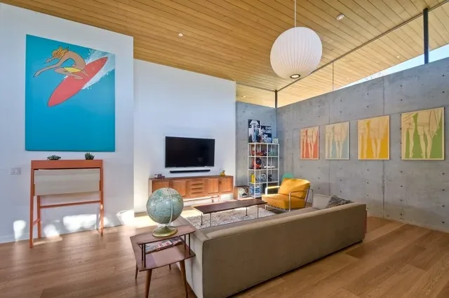Living room in the Aa House by Lloyd Russell AIA features a wooden ceiling, concrete walls, and midcentury modern furniture with colorful artwork.