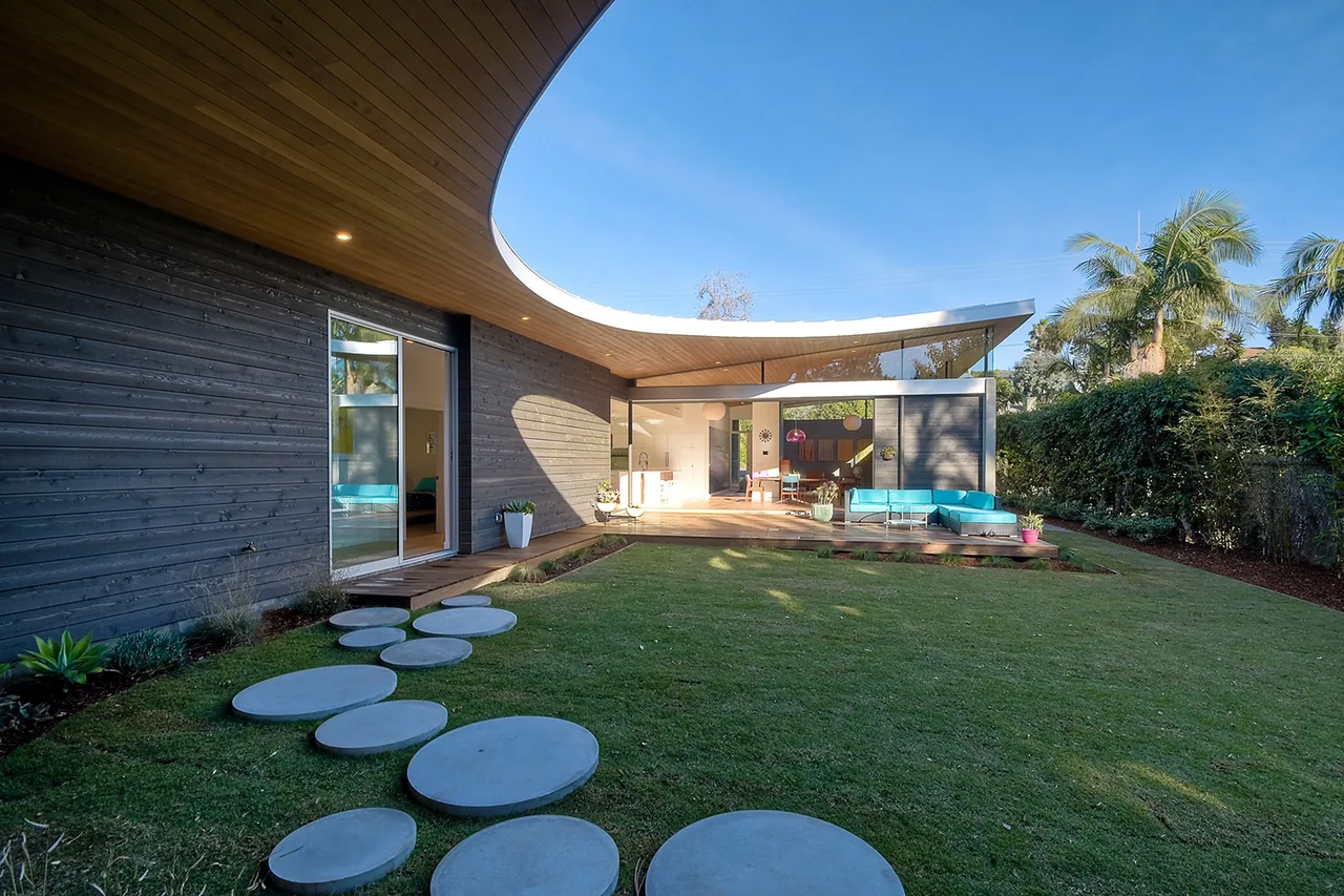 Modern house exterior features black wood siding, a curved wood ceiling overhang, and a backyard with round stepping stones leading to a patio.