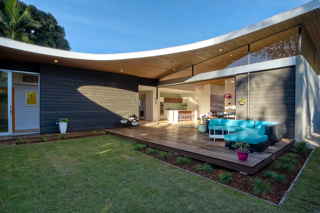 Modern exterior view of Aa House featuring a curved flat roof, wood siding, and an outdoor deck with seating and plants.