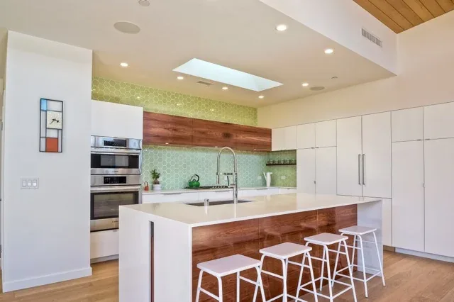Modern kitchen featuring a white island with a wooden base, green tile backsplash, skylight, and built-in stainless steel appliances.