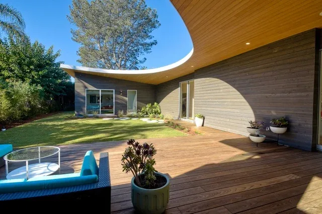 Deck area of Aa House featuring a curved wood ceiling overhang and a grassy backyard with round concrete stepping stones.