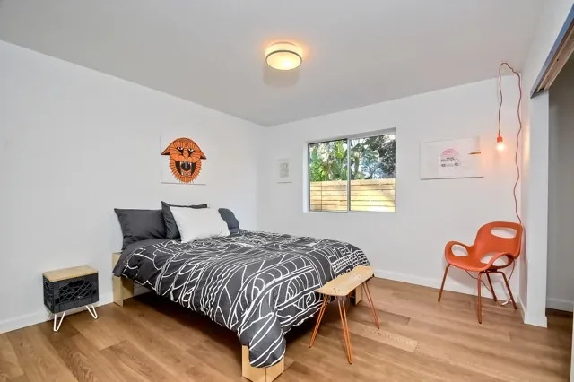 Minimal bedroom with wood flooring, a bed with black and white bedding, and a vibrant orange chair by the window.
