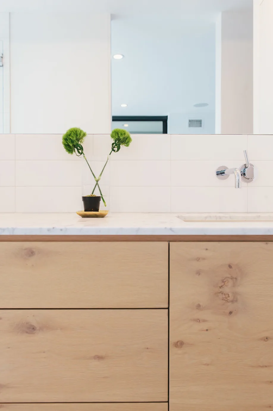 Minimalist bathroom vanity with light wood cabinetry, marble countertop, wall-mounted faucet, and a small decorative vase.