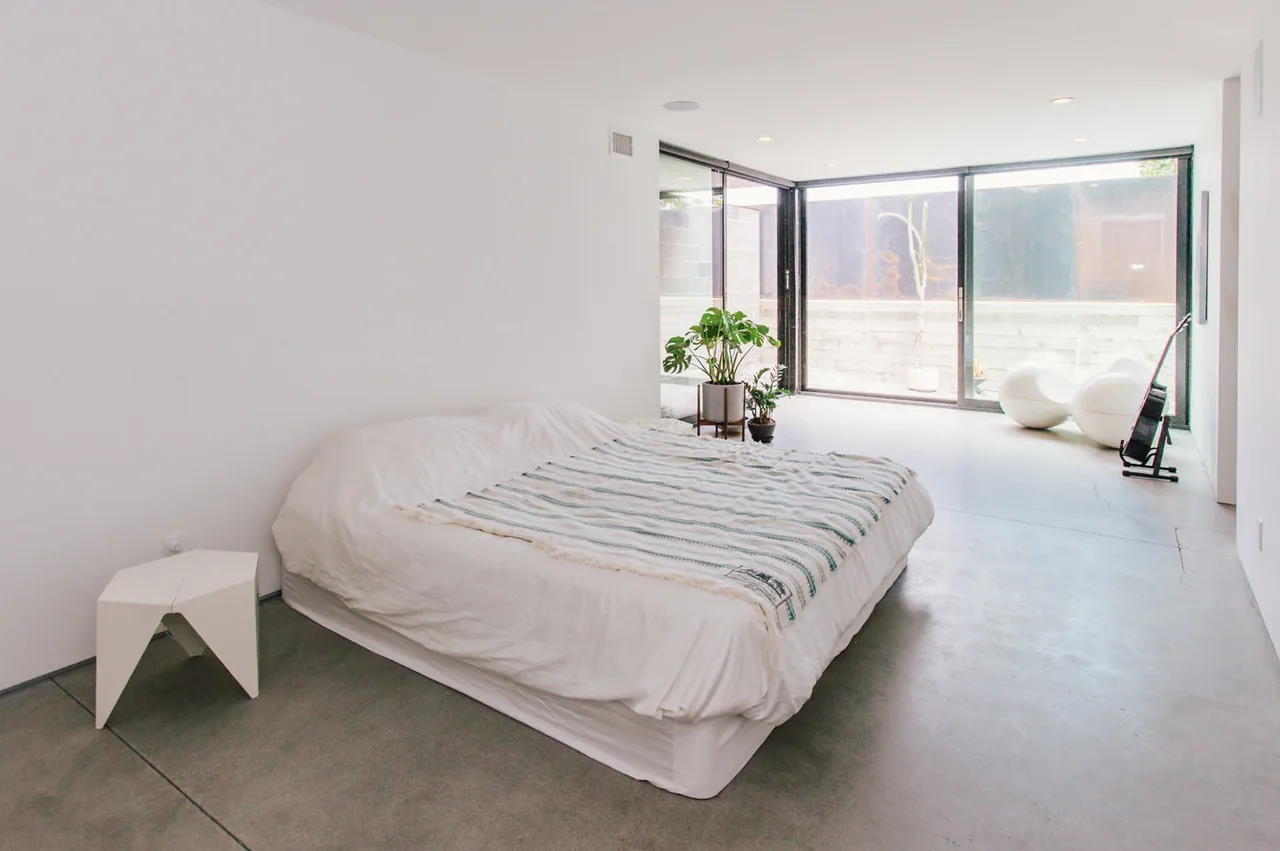 This minimalist bedroom features a low bed and large sliding corner windows providing natural light and outdoor views.