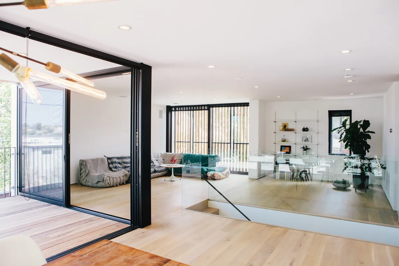Living area featuring light wood flooring, black-framed sliding glass doors, and minimal furniture with shelving and plants.