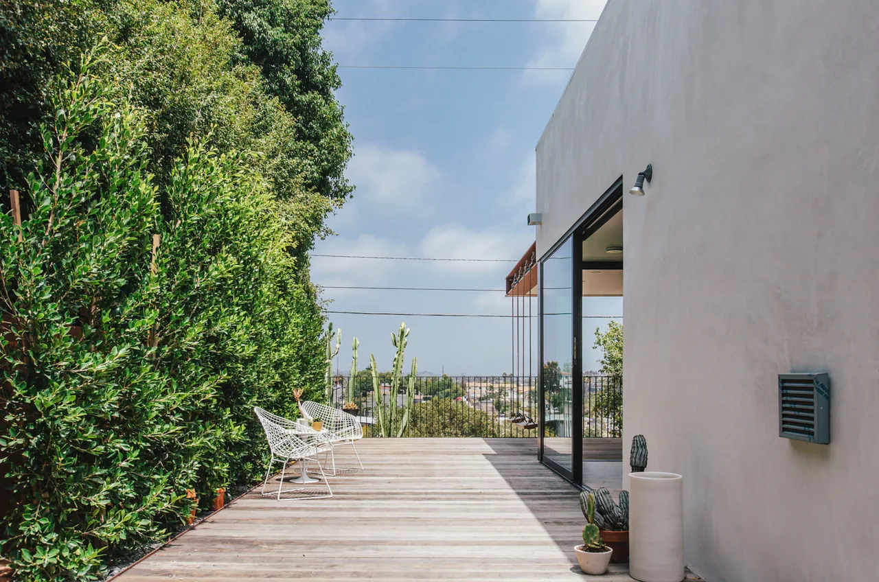Wooden deck patio with white wire chairs and potted cacti beside a modern building with sliding glass door.