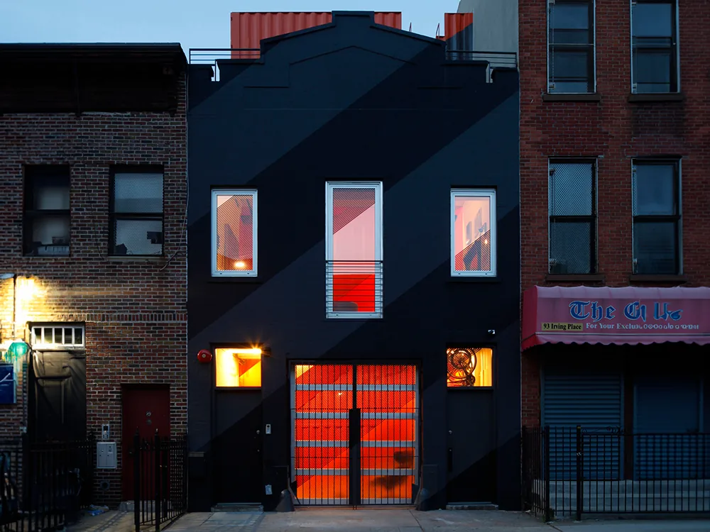 The Irving Place Carriage House features a black facade with diagonal stripes and glowing orange interior lighting at night.