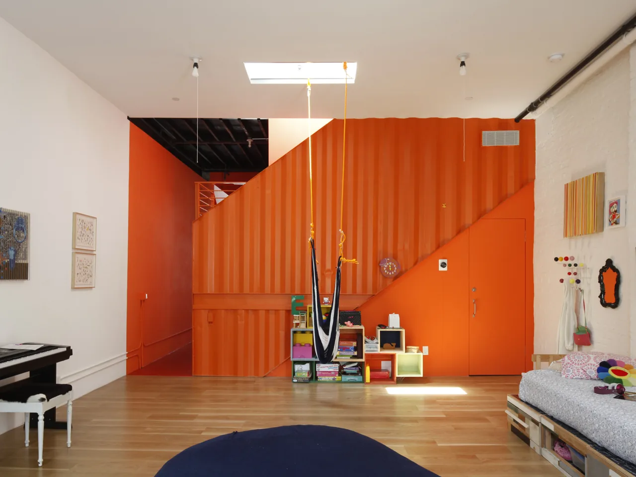 Room features an orange corrugated accent wall with an integrated door, wooden flooring, and a hammock chair under a skylight.