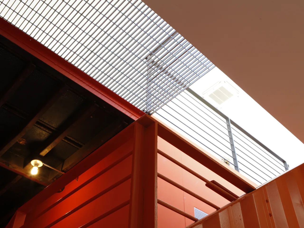 Interior stairwell featuring orange metal railings and a grated metal ceiling allowing natural light.