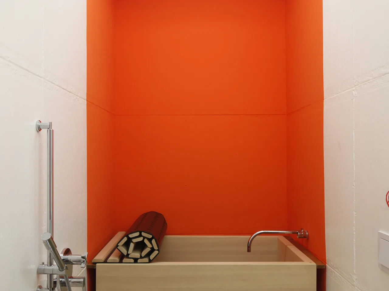 Rectangular wooden soaking tub set against a vibrant orange accent wall in a minimalist bathroom.