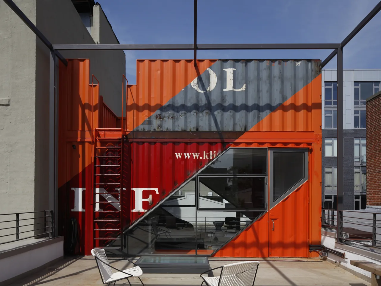 Shipping container structure with orange paint, metal stairs, and large diagonal glass windows forms an exterior design element.
