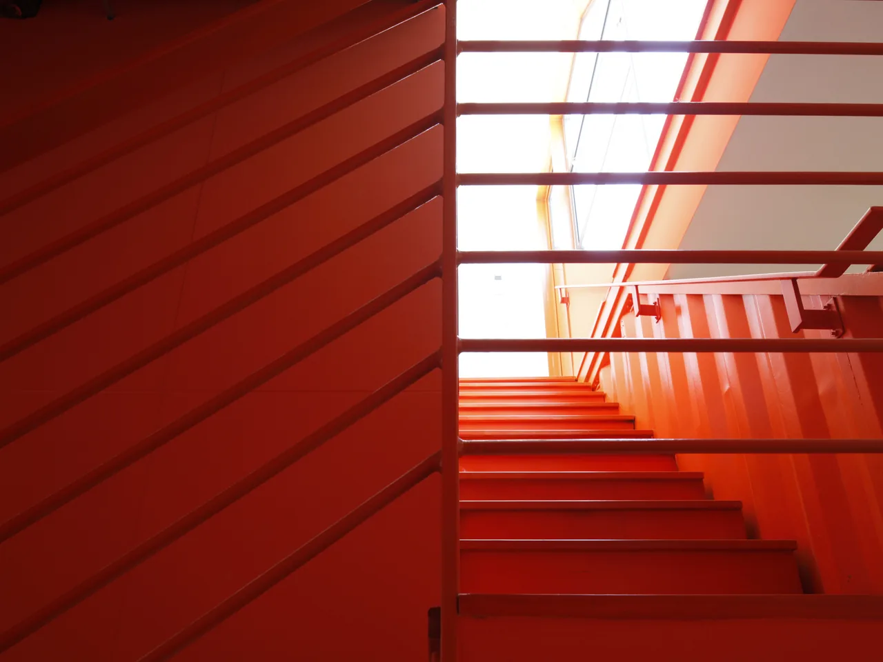 The photograph shows an orange-painted metal staircase with horizontal railings and a window above providing natural light.