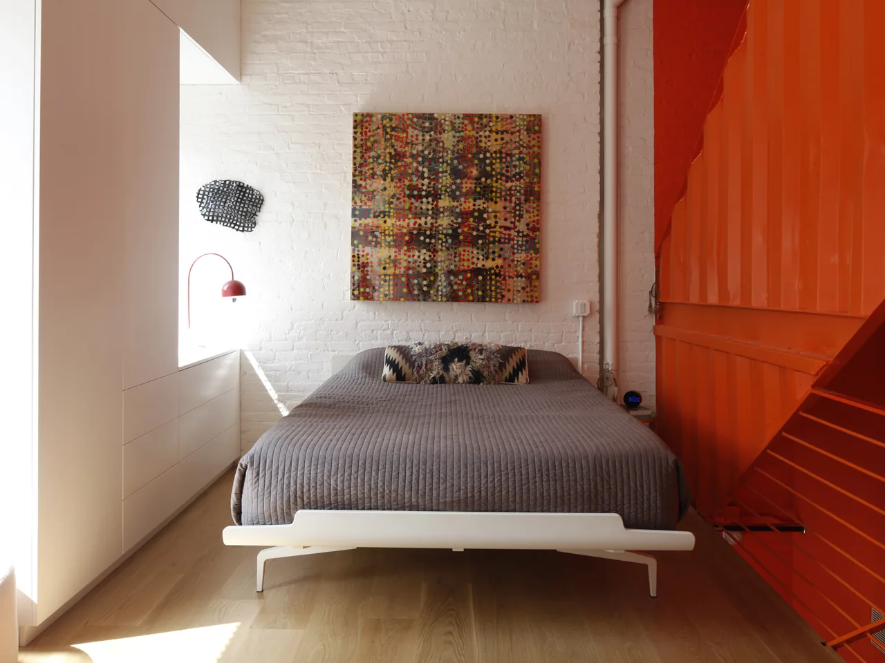Bedroom with gray bedding, abstract wall art, painted white brick wall, and an orange metal staircase wall.