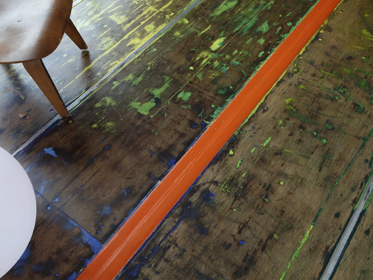 Floor detail showing dark wood with colorful paint splatters and an orange painted central strip near a wooden chair leg.