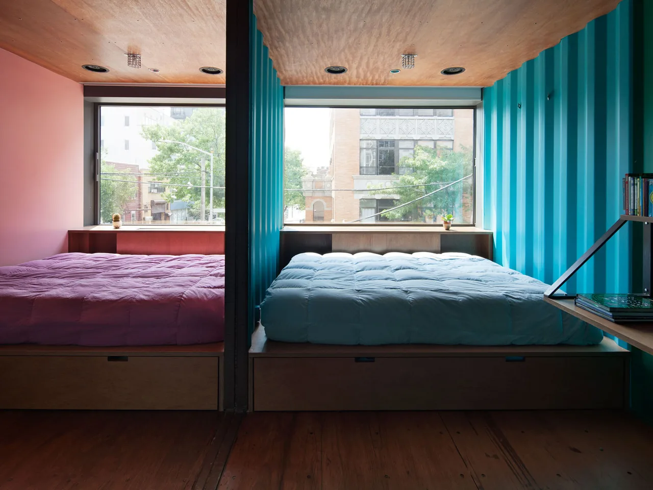 Bedroom with two beds separated by a blue partition wall and large windows showing urban views.