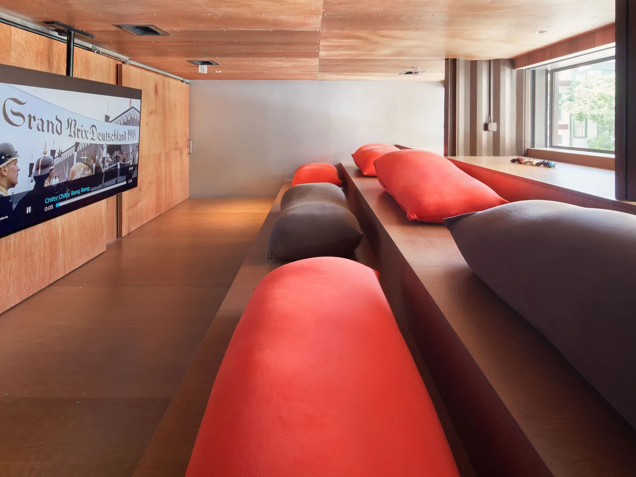 Tiered wooden bench seating area with orange and gray cushions and a wall-mounted TV in the CARROLL HOUSE media room.