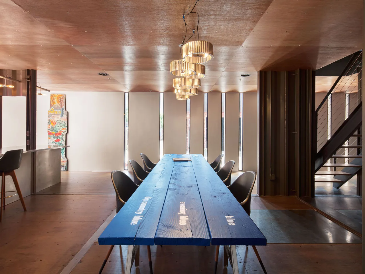 Dining space with vertical slit windows, a blue wooden table, and visible metal staircase in an industrial style interior.