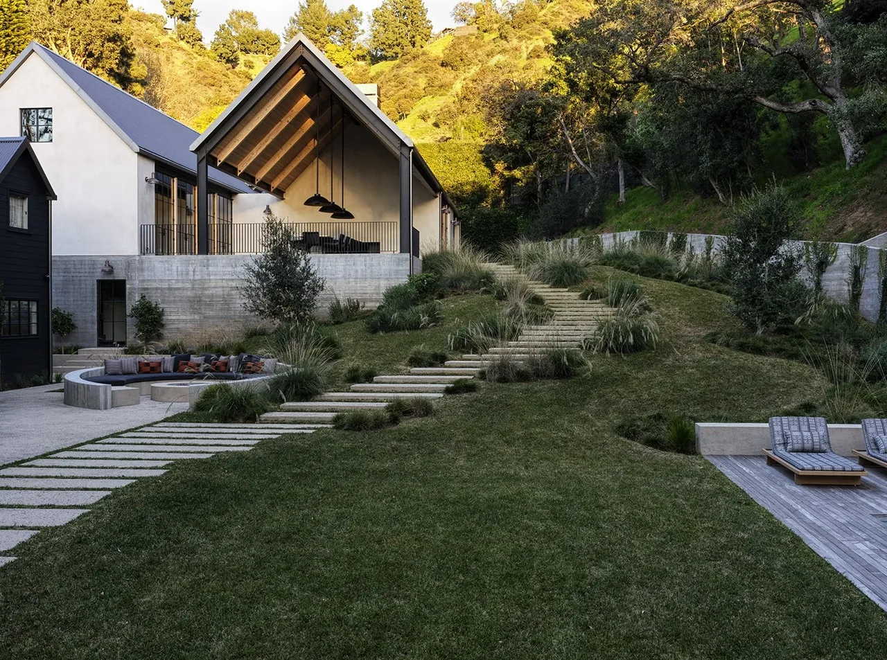 Canyon House backyard features a sloped lawn with concrete steps and outdoor seating beside a house with gable roofs and a covered patio.