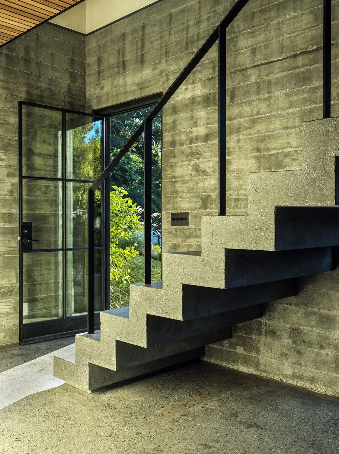 Concrete stairs with black metal railing beside large glass door in a textured concrete wall interior.