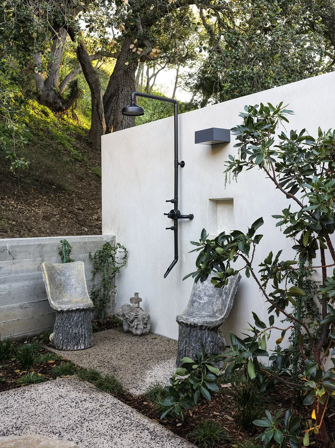 Outdoor shower space with black metal shower fixture on white stucco wall, stone chairs, and lush landscaping creating a natural retreat.