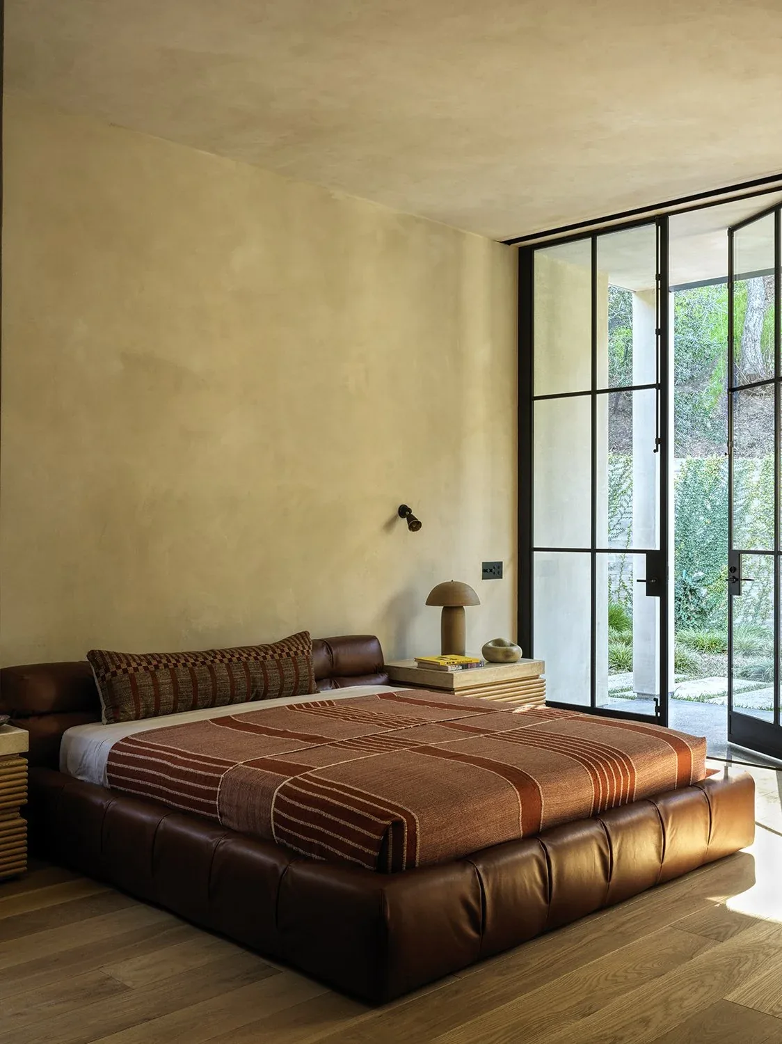 Bedroom featuring a brown leather bed frame, textured walls, and floor-to-ceiling black framed glass doors leading outside.